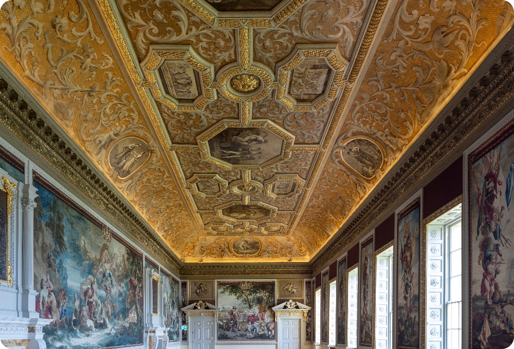 A large rectangular dining room a huge gold glittering ceiling covered in paintings and decorative scrolls with octangular shaped spaces for the paintings. The room on the right has five windows and in between each is a different scene of wall trophies displayed on wallpaper to resemble the original tapestries. The paint in between is also a maroon colour. On the left hand side of the room is a very large scene titled the triumph of Venus which depicts a Grecian myth to resemble the original tapestry and is made of wallpaper. On either side of it are two large fireplaces with over Mantel carvings. At the far end of the room are two large doors, Intricately carved and above each is a painting of a centaur in grisailles format. Another scene is depicted on a tapestry wallpaper titled The Triumph of Ceras, the goddess of harvest sitting in her chariot being drawn by leopards. The room is intricately decorated with lots of gold gilding and is at Stowe House in Buckinghamshire.