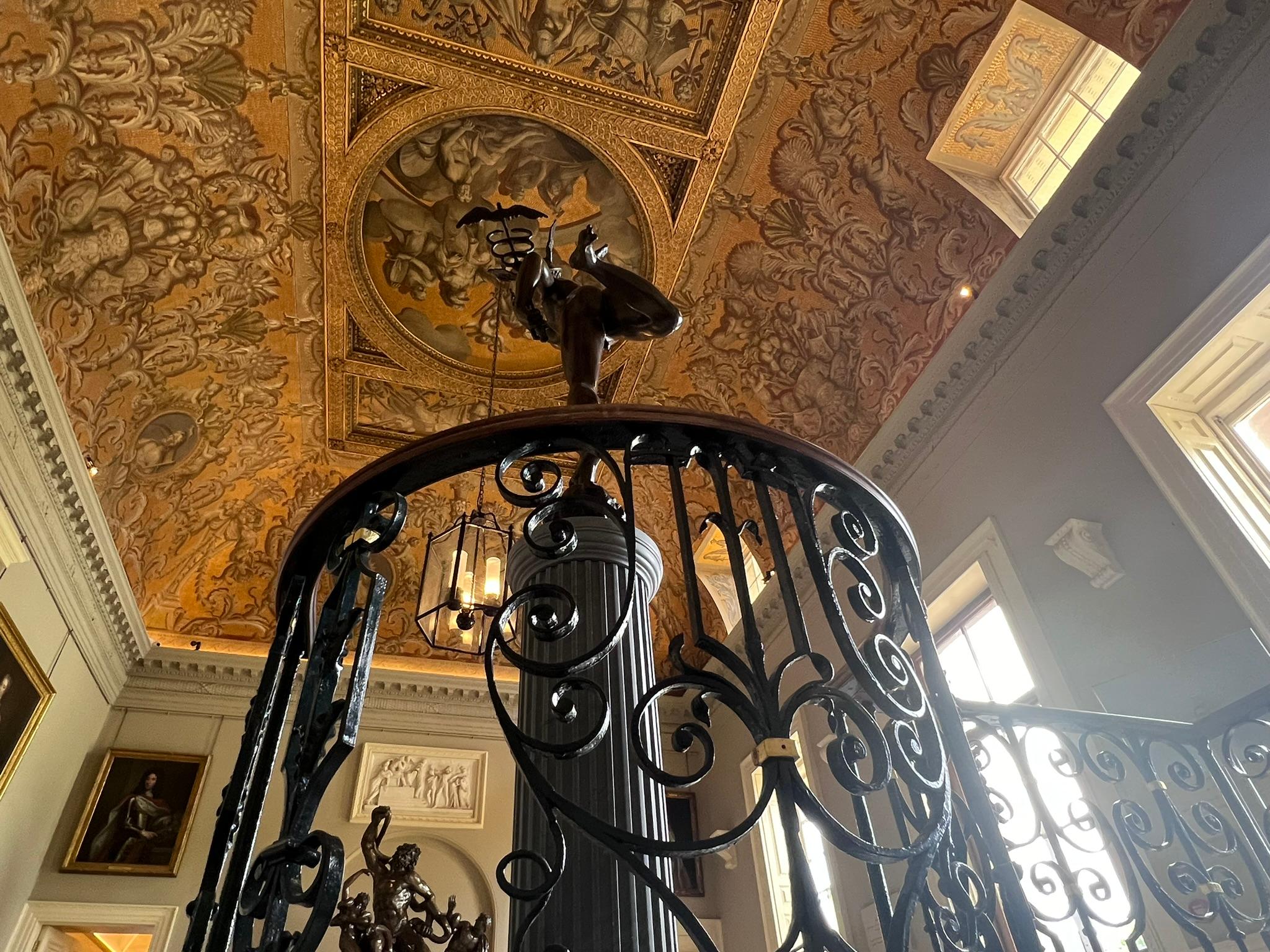 An entrance room with large wooden doors, twice human height with glass panels to enter. The ceiling is glowing gold and covered in paintings by William Kent showing roman gods to depict the months and astronomical symbols. A large bronze statue of the mercury stands in the room. There is a large lantern hanging from the ceiling and a doorway leading to a corridor. The view looks up from a curved stairwell