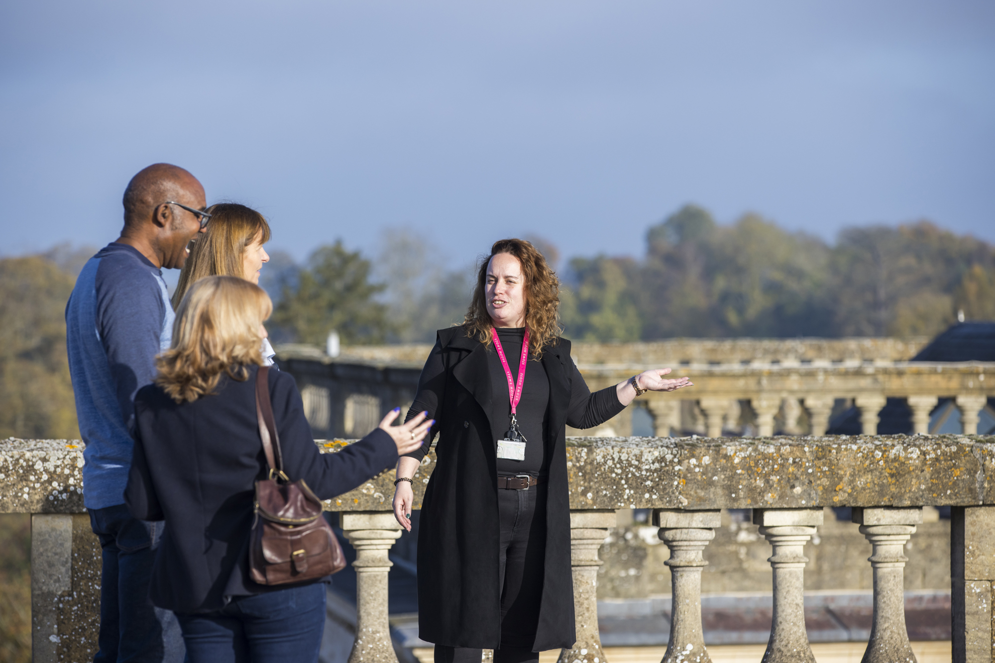 Four people chatting on a sunny day during a rooftop tour, standing by a stone balustrade with trees ain the background.