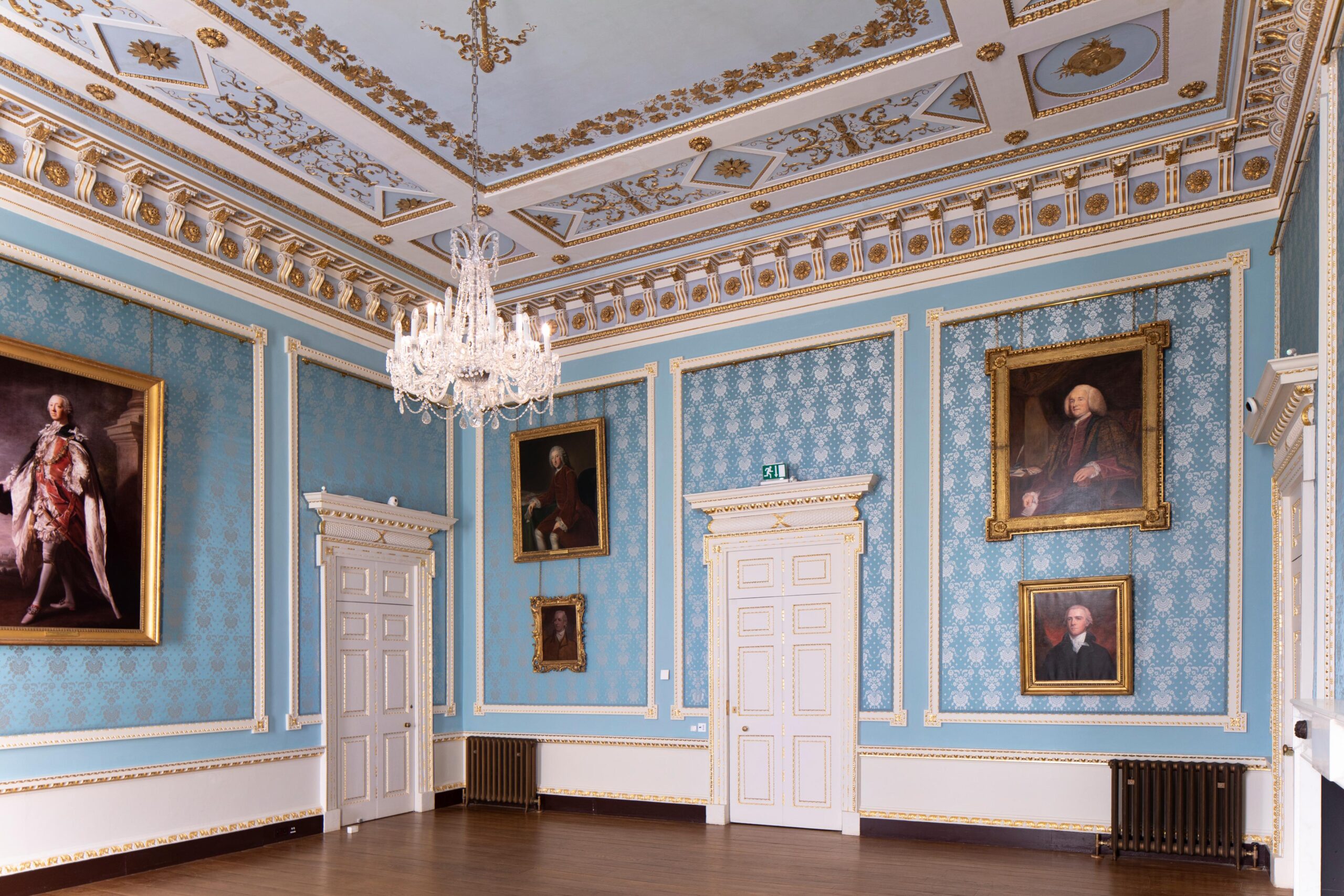 A square room which is covered in blue silk damask wallpaper. the ceiling is blue and white with wooden intricate carvings and gold gilding. the floor is oak. a chandelier hangs from the ceiling. Portraits of prime ministers related to the Temple-Grenville family at Stowe hang on the walls.