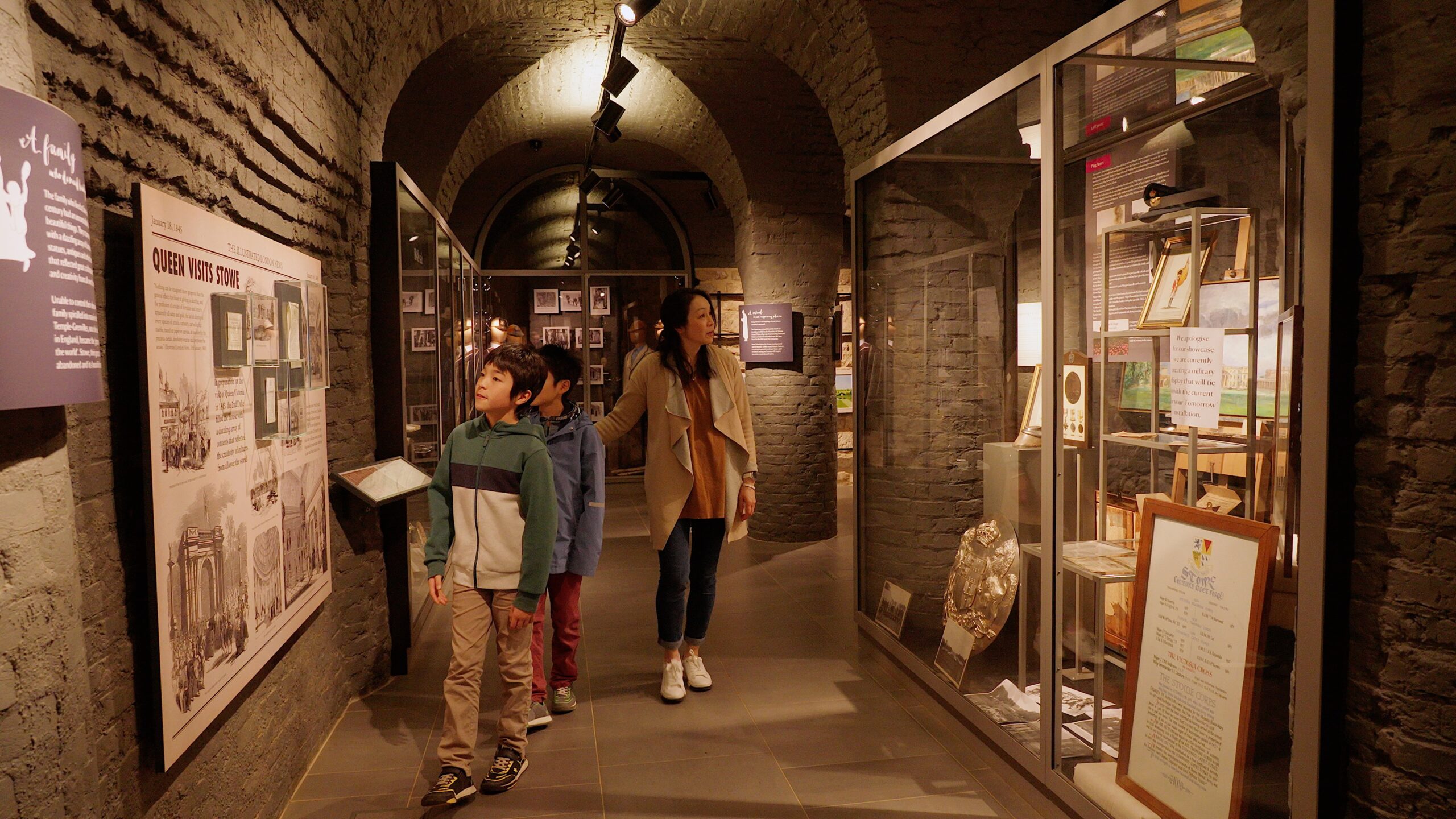 A Japanese mum in her forties and her two young sons walk through the museum at Stowe House. The museum is in an old wine cellar with a vaulted ceiling and the brick work is grey. there are information boards on the walls and glass display cabinets either side of them.