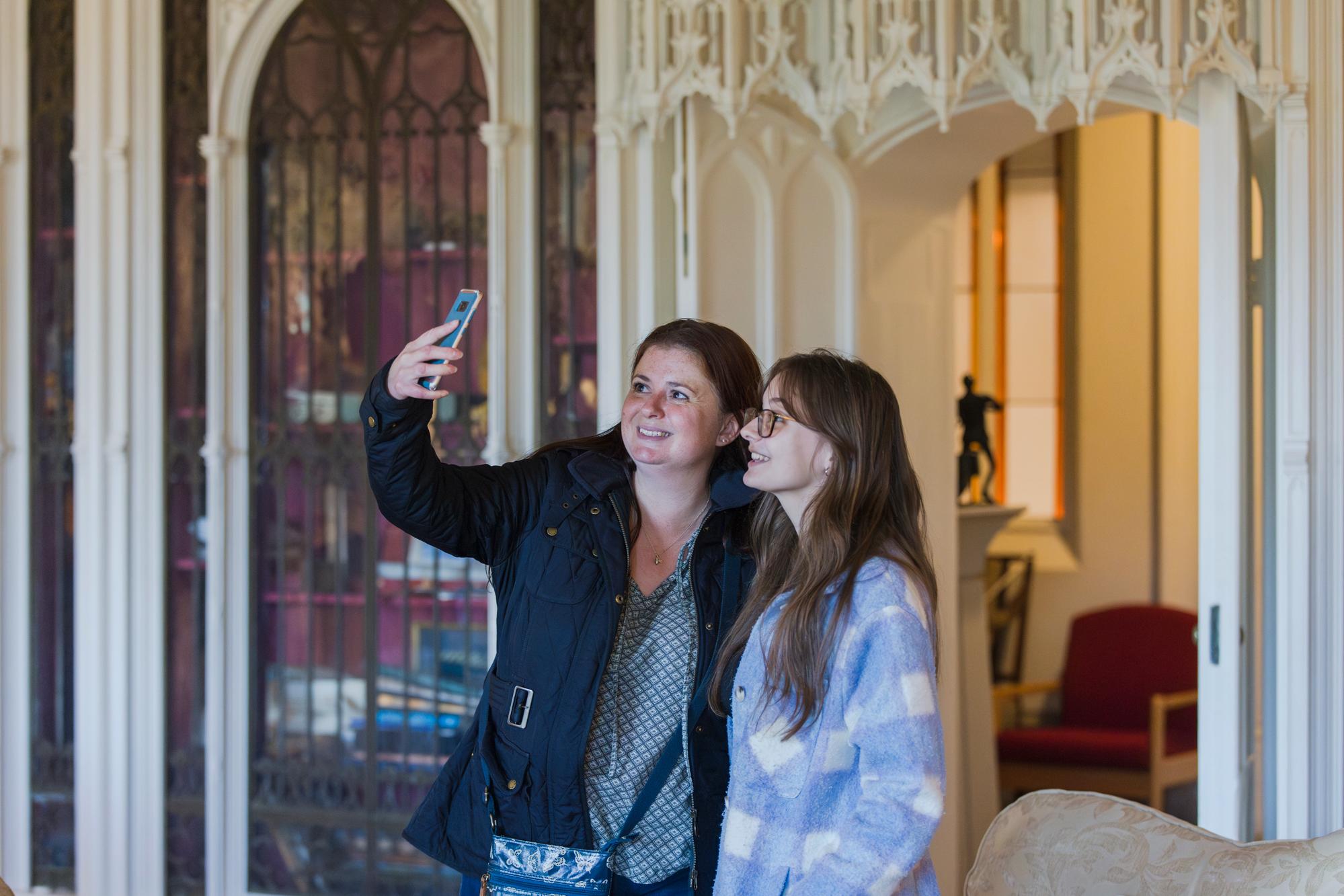 A white woman in her forties with long cherry brown colour hair, wearing a coat has her arm around a young teenage white girl with long brown hair wearing glasses. the both smile and the women holds up her phone to take a selfie photo. they stand in a decorated room of gothic style