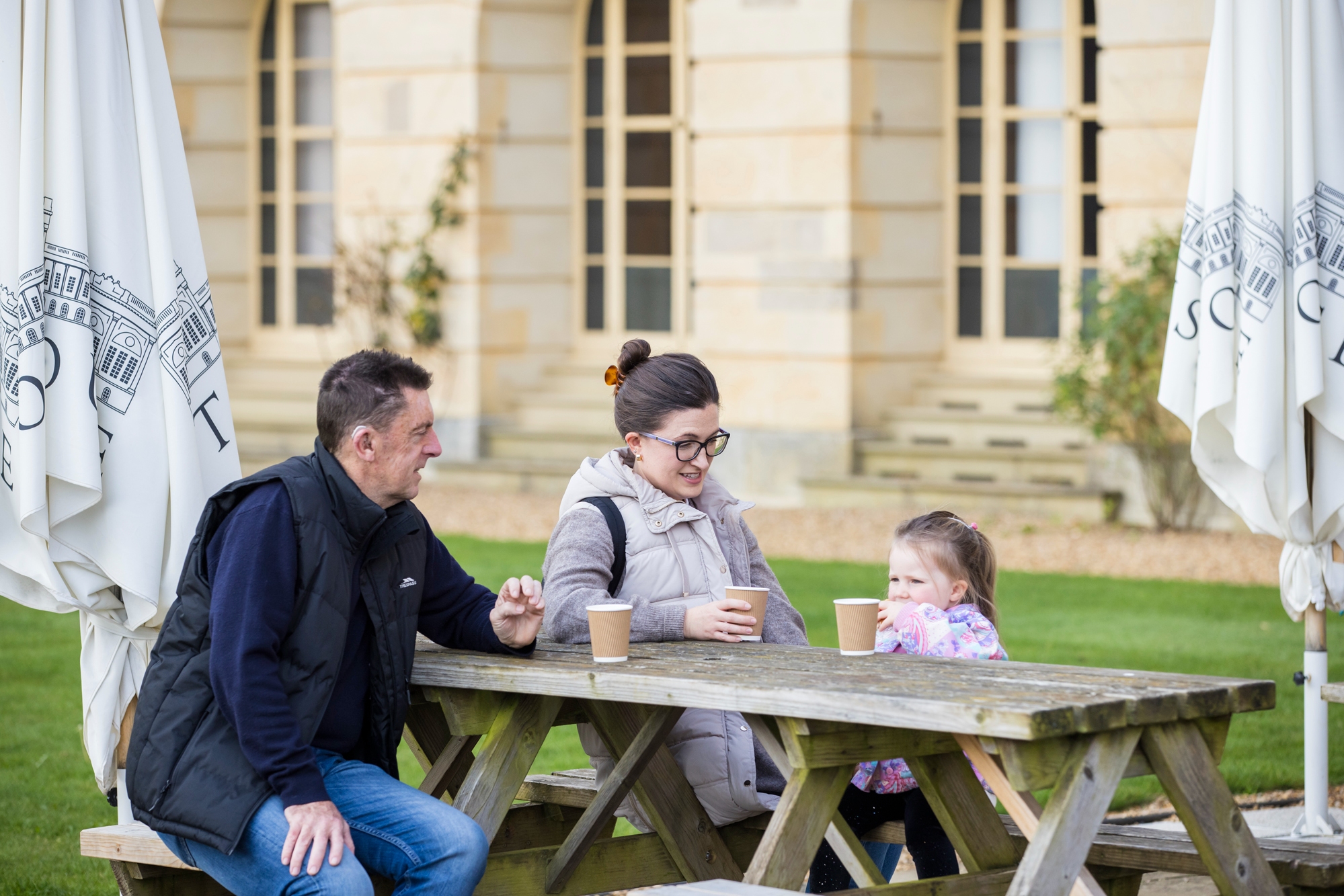 A family consiting of a grandad, daughter and grandaughter sit on a picnic bench at stowe house