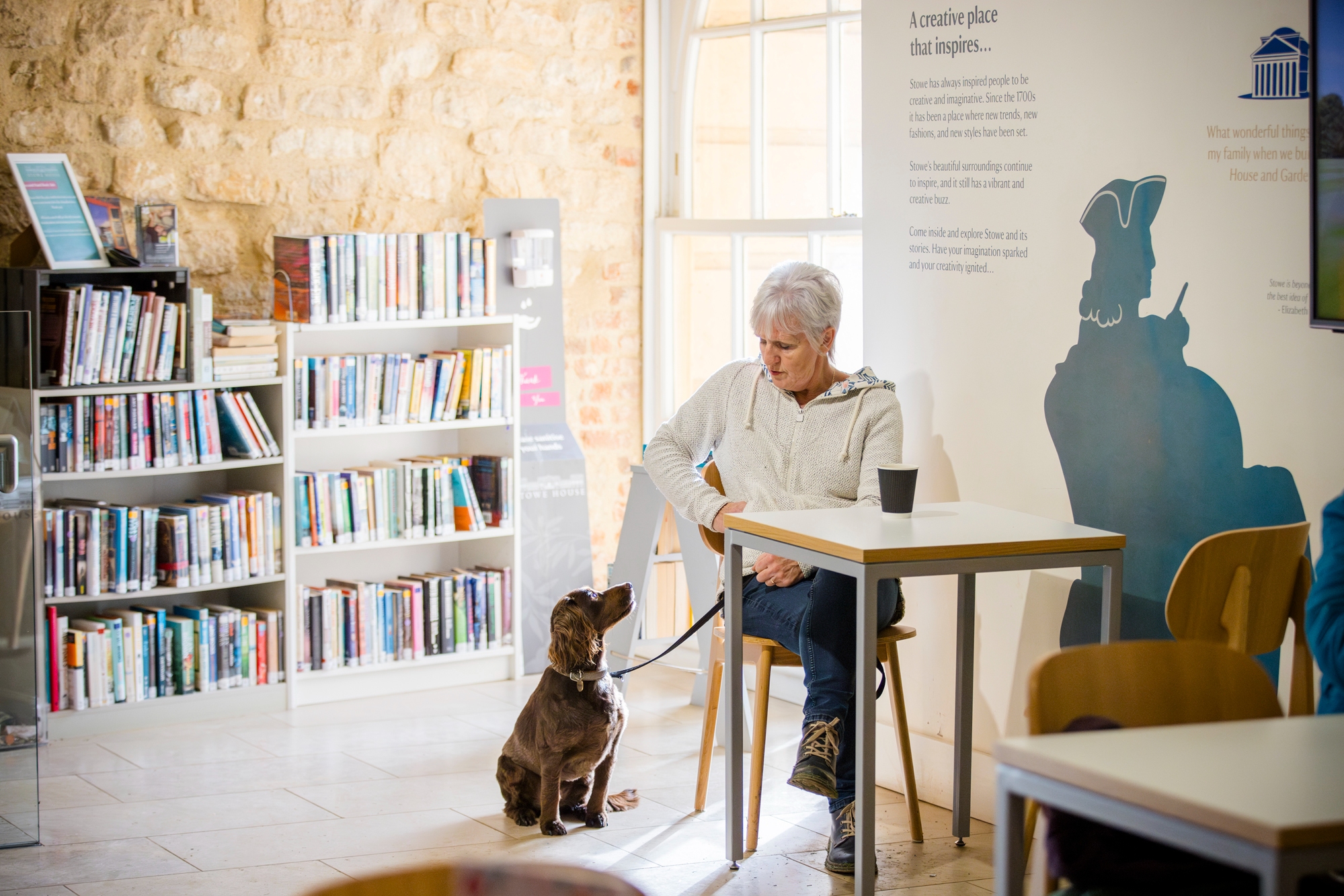 A woman in her 60s sits at a table with a dog sitting on the floor next to her on a lead in the visitor centre at Stowe House