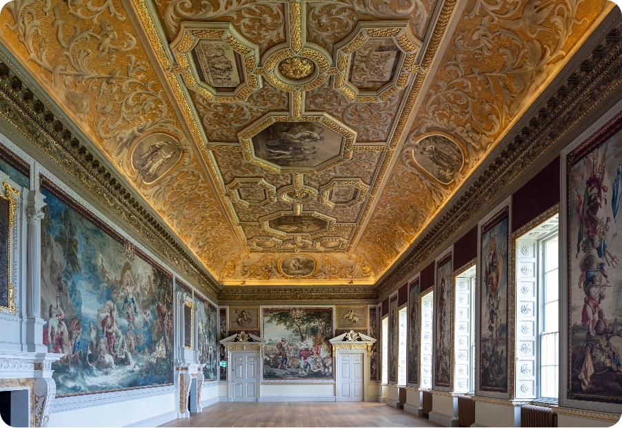 A large rectangular room with no furniture. There are oak floorboards, a ceiling which glows gold and is covered in intrictae paintings from the eighteenth century, some of greek and roman myths, others of animals. The walls are covered with replica wallpaper of what was once original tapestries displaying scenes from roman myths. On the left are two fireplaces with tall overmantles. the dados, door frames, and wooden elements in the room are all gilded with gold. Bright sunlight comes through the many tall rectangular windows on the right side of the room.