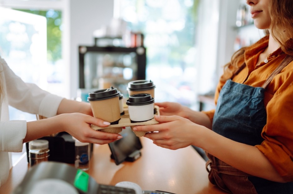 Two women stand and are passing between them hot drinks to go.